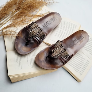 May include: A pair of brown leather sandals with a beaded strap and a woven design. The sandals are on a book with a white background.