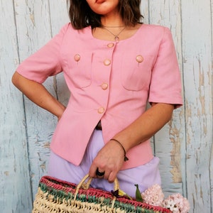May include: A pink short-sleeved button-up blouse with gold buttons and two flap pockets. The blouse is worn with a light purple skirt and a woven straw bag with colorful accents.