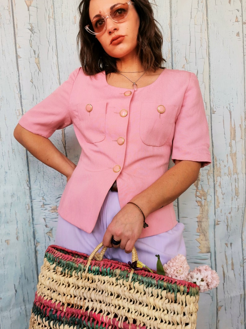 May include: A pink short-sleeved button-up blouse with gold buttons and two flap pockets. The blouse is worn with a light purple skirt and a woven straw bag with colorful accents.
