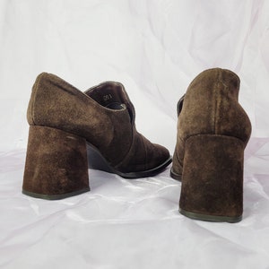 May include: A pair of brown suede heeled ankle boots with a block heel. The boots have a pointed toe and a zipper on the inside of the ankle.