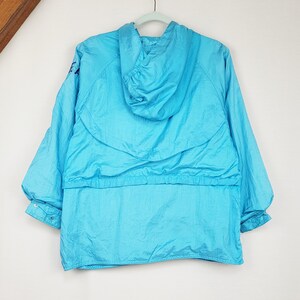 May include: A bright turquoise hooded jacket hangs on a light green hanger. The jacket has a drawstring waist and elastic cuffs. The material has a slight sheen. The jacket is a vintage style.