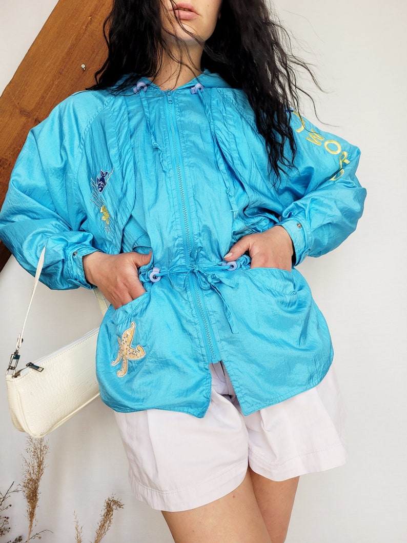 May include: A turquoise vintage windbreaker jacket with a hood, zipper, and drawstring waist. The jacket features embroidered floral and starfish designs. The sleeve has the word "SPORT" in yellow. The jacket is paired with white shorts.