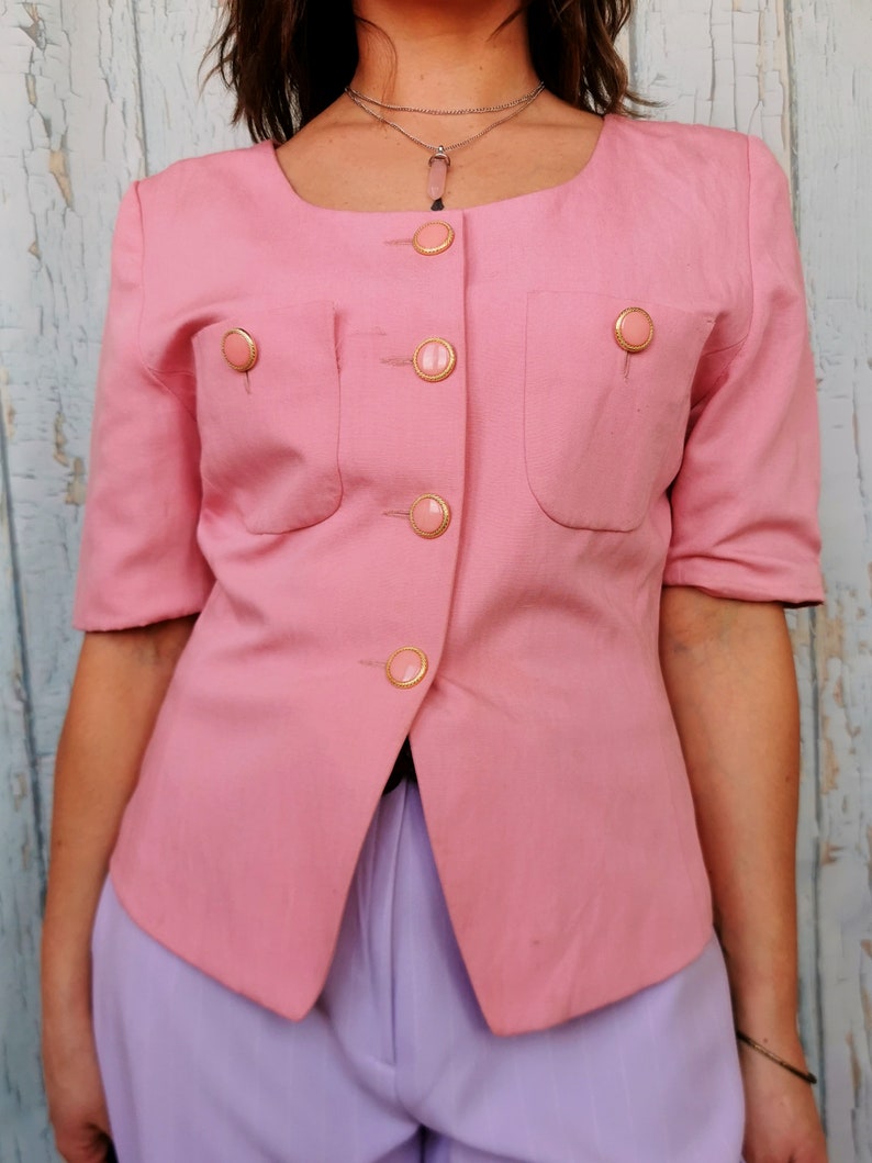 May include: A pink blazer with gold buttons and a scoop neck. The blazer has two pockets on the front and is made of a lightweight fabric.