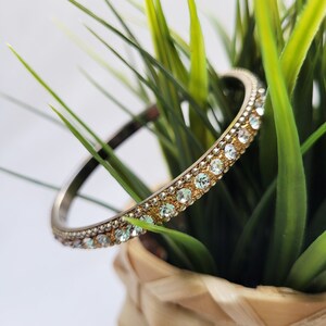 Könnte beinhalten: Ein goldenes Armreif-Armband mit klaren Strasssteinen, die in eine silberne Metallband gefasst sind.