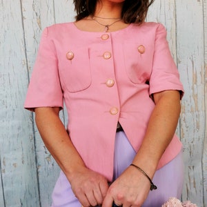 May include: A pink short-sleeved button-up blouse with gold buttons and a single pocket on the front. The blouse is worn with a light purple skirt and a woven straw bag.