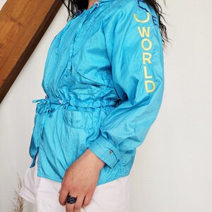 May include: A turquoise blue windbreaker jacket with the words "SEE WORLD" embroidered in yellow on the sleeve. The jacket has a drawstring waist and a hood. The person is wearing white shorts.