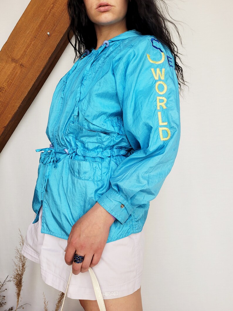 May include: A turquoise blue windbreaker jacket with the words "SEE WORLD" embroidered in yellow on the sleeve. The jacket has a drawstring waist and a hood. The person is wearing white shorts.