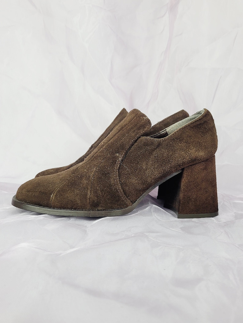 May include: Brown suede heeled shoes with a block heel. The shoes have a pointed toe and a slip-on design.