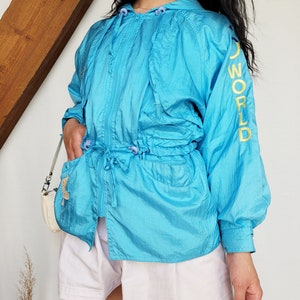 May include: A turquoise hooded jacket with a drawstring waist and zipper closure. The jacket has the word "WORLD" in yellow lettering on the sleeve. The model is wearing white shorts and a small white purse.