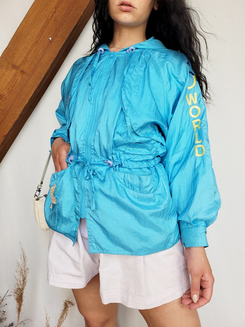 May include: A turquoise hooded jacket with a drawstring waist and zipper closure. The jacket has the word "WORLD" in yellow lettering on the sleeve. The model is wearing white shorts and a small white purse.