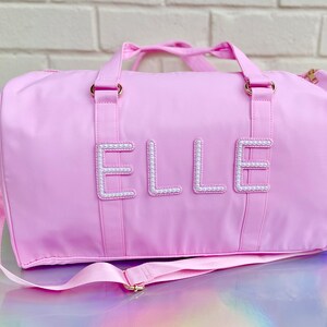 Peut inclure: Un sac de sport rose avec un motif "ELLE" orné de perles blanches. Le sac a une longue sangle et deux poignées supérieures.