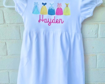 Könnte beinhalten: Weißes Kleid mit kurzen Ärmeln und einem Rüschen-Saum. Das Kleid hat ein gesticktes Design mit fünf Prinzessinnenkleidern und dem Namen "Hayden" in Rosa.