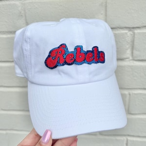 May include: White baseball cap with the word "Rebels" embroidered in red with a blue outline. The cap has a curved brim and a classic, casual style. The hat is made of a soft, durable material.