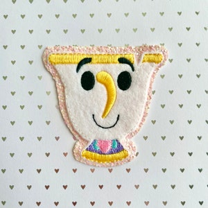May include: A felt applique of a cartoon teacup character with a smiling face. The teacup is white with a yellow nose and rim, black eyes and eyebrows, and a colorful collar. The applique is outlined with pink glitter.