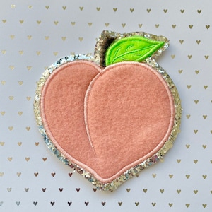Könnte beinhalten: Ein pfirsichförmiger Filz-Patch mit einem grünen Filzblatt, umgeben von einem silbernen Glitzerbesatz.