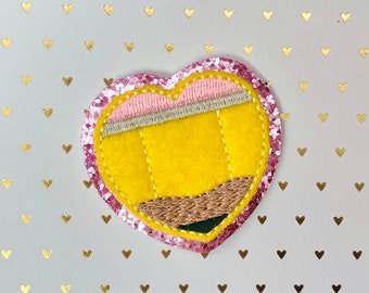 May include: A pink heart-shaped felt patch with a yellow pencil design embroidered on it. The pencil has a brown eraser and a pink and white striped band.