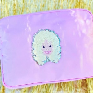 Puede incluir: Un estuche rectangular de color rosa claro con cremallera que presenta un parche bordado central de una persona con cabello rubio voluminoso y rostro sonriente. El parche tiene un borde de purpurina plateada y la superficie es lisa.