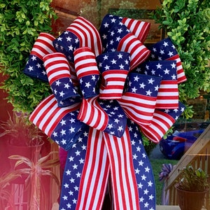 May include: A large red, white, and blue bow made of fabric with a stars and stripes pattern. The bow is tied in a large loop and is perfect for decorating for a patriotic holiday.