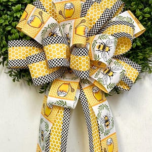 May include: A yellow and black checkered ribbon with a white background featuring a repeating pattern of honey jars, bees, and green leaves. The ribbon is tied into a bow.