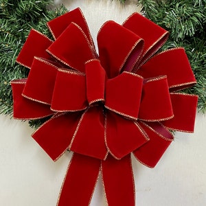 May include: A large, decorative bow made of deep red velvet ribbon with gold trim. The bow is attached to a green garland, creating a festive holiday decoration. The bow's loops and tails are prominently displayed.