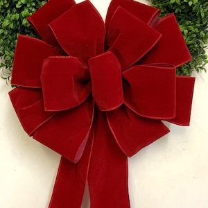 May include: A large, vibrant red velvet bow with multiple loops and long tails. The bow is set against a backdrop of green foliage, creating a festive contrast. Perfect for holiday decorations or gift wrapping.