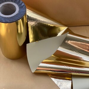 Op de afbeelding: Een rol gouden metallic inpakpapier met een witte achterkant. Het papier is opgerold en het uiteinde is omgevouwen. Het papier is glanzend en reflecterend.