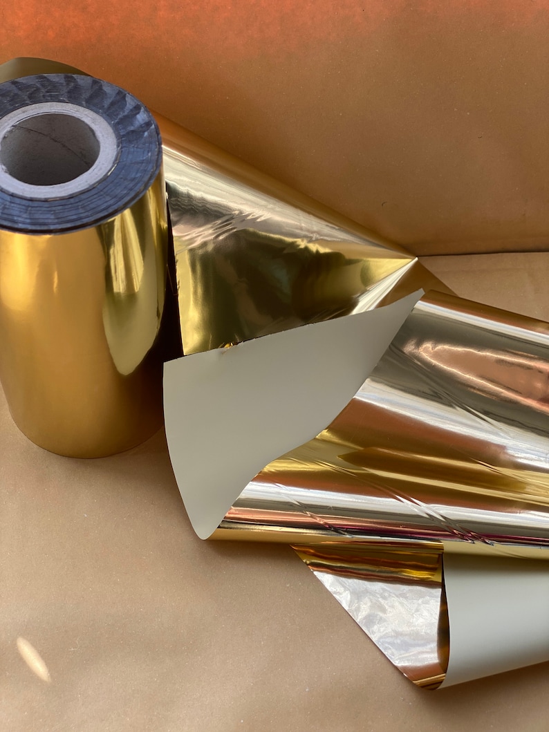 Op de afbeelding: Een rol gouden metallic inpakpapier met een witte achterkant. Het papier is opgerold en het uiteinde is omgevouwen. Het papier is glanzend en reflecterend.
