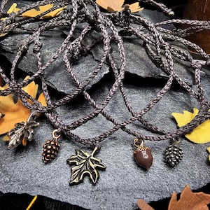 May include: Braided cord bracelets with autumn-themed charms. The charms include acorns, leaves, and pinecones in bronze, silver, and brown tones. The bracelets are arranged on a dark gray surface with fall foliage accents.