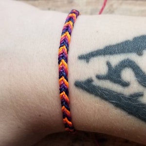May include: Braided bracelet in shades of purple, orange, and black. The bracelet is worn on a wrist. The bracelet is made of woven threads. The bracelet is a handmade accessory.