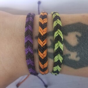 May include: Three braided cord bracelets featuring a herringbone pattern. One bracelet is purple and black, the second is orange and black, and the third is lime green and black. Each bracelet is constructed from thin, twisted cord material.