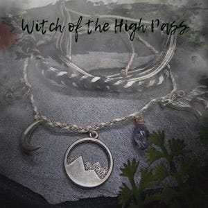 May include: A collection of handmade bracelets and necklaces with a mystical theme. The jewelry features silver-toned charms, including a crescent moon, a mountain scene, and a wolf. The text "Witch of the High Pass" is written in cursive at the top.