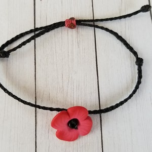 May include: Black cord bracelet featuring a red poppy flower with a black center. The adjustable bracelet is tied with knots and has a small red accent bead. The poppy is a symbol of remembrance.