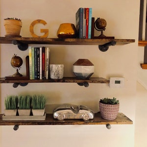 May include: Three wooden shelves with black metal brackets, decorated with a variety of items, including books, globes, plants, and decorative objects. The shelves are mounted on a white wall.