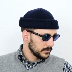 Puede incluir: Un hombre con un gorro azul, gafas de sol de color azul, un chaleco de punto gris con un pequeño logotipo y una camisa a cuadros.