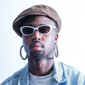 May include: A person wearing a white t-shirt, a blue denim jacket, a brown and white patterned newsboy cap, white oval sunglasses, and large silver hoop earrings. The person has a nose ring.