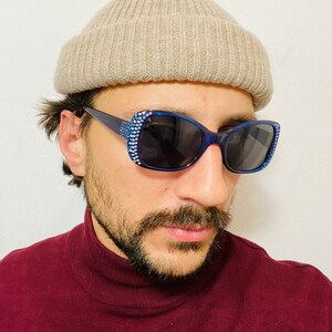 Puede incluir: Una persona con una boina beige y gafas de sol azules con pedrer&iacute;a. Las gafas tienen lentes oscuras. La persona lleva un jersey de cuello alto marr&oacute;n.