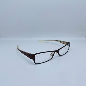 May include: Brown rectangular eyeglasses with clear lenses. The frames are a dark brown color with a subtle design on the temples. The arms of the glasses are a lighter color with a striped pattern. The glasses are set against a white background.