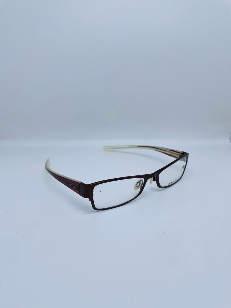 May include: Brown rectangular eyeglasses with clear lenses. The frames are a dark brown color with a subtle design on the temples. The arms of the glasses are a lighter color with a striped pattern. The glasses are set against a white background.