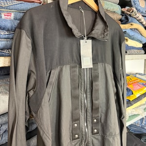 Puede incluir: Chaqueta negra de dos tonos con cremallera y cuello alzado. La etiqueta está adjunta y la chaqueta está colgada de una percha de madera. La chaqueta parece estar hecha de un material duradero, posiblemente resistente al agua.