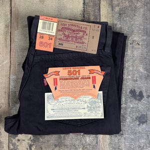 May include: Black Levi's 501 jeans with original tags attached. The tags display the Levi Strauss & Co. logo, the 501 model name, and size specifications of 28 inch waist and 34 inch inseam. The jeans are made of 100% cotton and feature a button-fly.