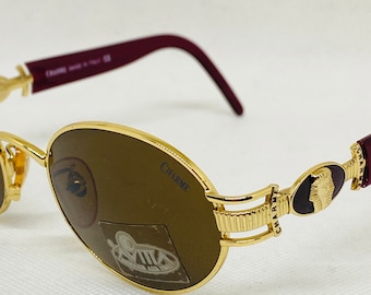 May include: Gold-tone oval sunglasses with brown lenses and a burgundy frame. The sunglasses have a decorative gold accent on the side with the word "CHARME" written on the frame.