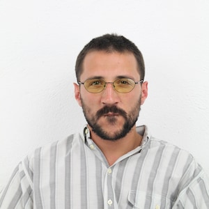 May include: A man with a beard wearing a striped button-down shirt and yellow-tinted glasses.