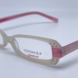 Könnte beinhalten: Weiße und rosafarbene Brille mit Strasssteinen an den Bügeln und dem Schriftzug "COCONUDA eyewear" auf dem Rahmen.