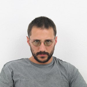 May include: A man with short brown hair and a beard is wearing round-rimmed glasses and a gray t-shirt. He is looking directly at the camera.