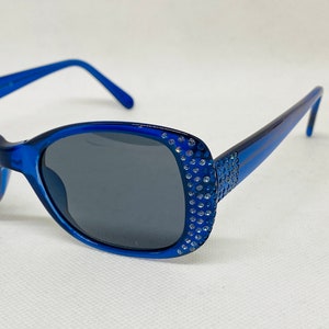 Puede incluir: Gafas de sol azules con un borde de strass alrededor de las lentes. Las gafas tienen lentes grises oscuras.