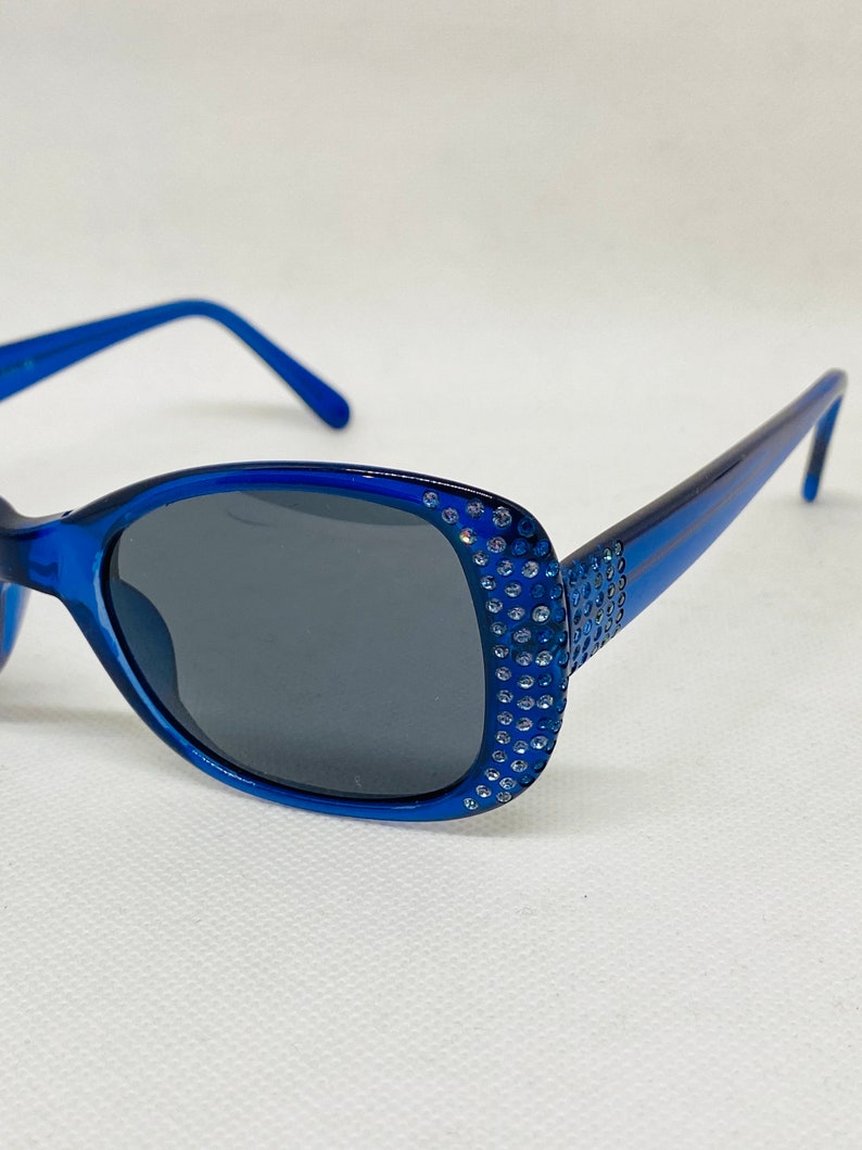 Puede incluir: Gafas de sol azules con un borde de strass alrededor de las lentes. Las gafas tienen lentes grises oscuras.