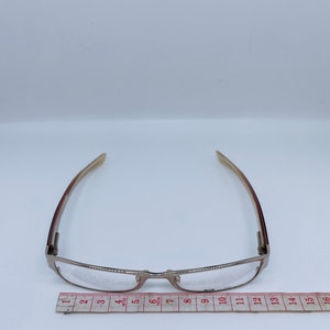 Może przedstawiać: Srebrne okulary do czytania z prostokątną oprawką i przezroczystymi szkłami. Okulary są mierzone na linijce, co pokazuje, że mają około 30 cm szerokości.