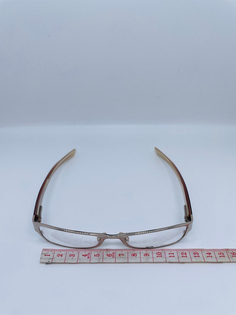 Może przedstawiać: Srebrne okulary do czytania z prostokątną oprawką i przezroczystymi szkłami. Okulary są mierzone na linijce, co pokazuje, że mają około 30 cm szerokości.