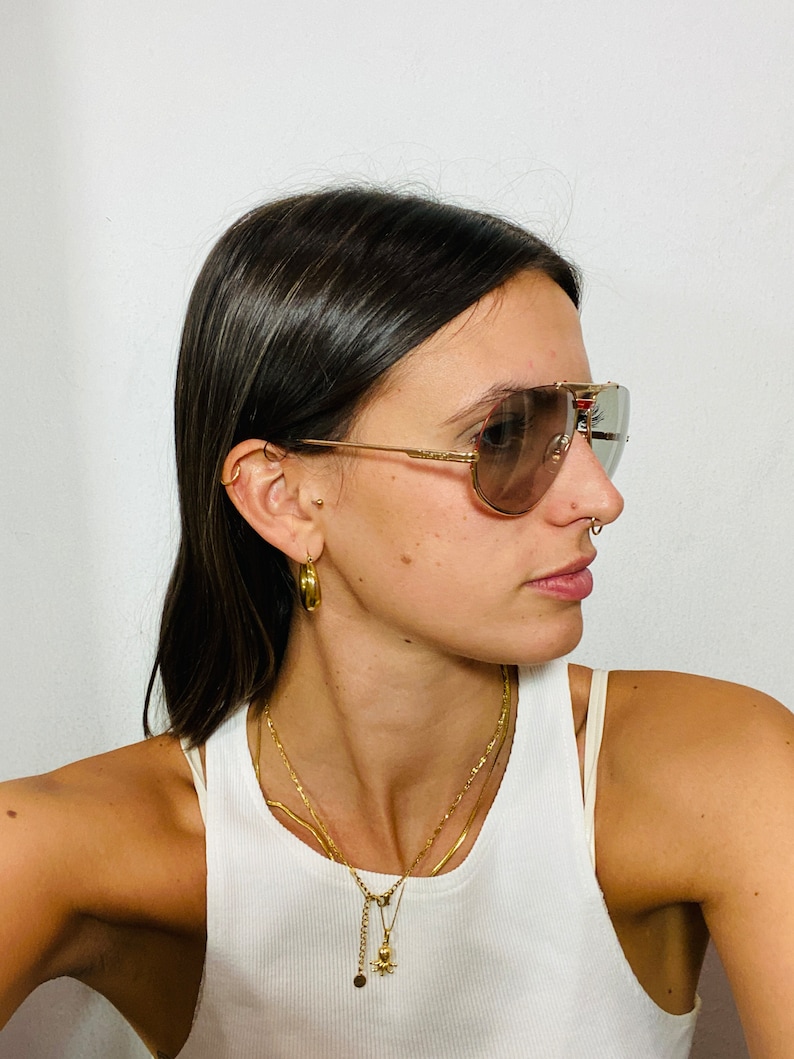 May include: A woman wearing gold hoop earrings, a gold chain necklace with a small octopus pendant, and a pair of brown aviator sunglasses with gold frames.