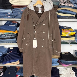 May include: A brown, double-breasted coat with a grey hooded lining. The coat features multiple silver buttons and a tag. It hangs on a wooden hanger. Shelves of folded jeans and other clothing form the background.